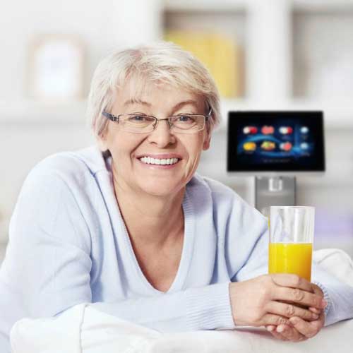 Lächelnde Seniorin mit einem Glas Orangensaft, im Hintergrund ein trinkForm® Getränkeautomat